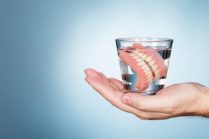 A hand holding a full set of dentures in a glass of clear liquid